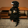 A person wearing a realistic dinosaur skeleton costume with a black headpiece and tail, posing in front of a wooden table.