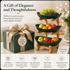 A gift box with a Best Wishes tag sits next to a 3-tier fruit basket stand filled with grapes, apples, and berries.