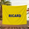 A bright yellow flag with the blue text RICARD is displayed outdoors against a backdrop of greenery and water.
