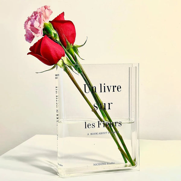 A clear acrylic book vase holds a red rose and a pink carnation.
