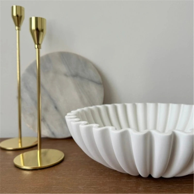 A white ruffled decorative bowl is displayed next to two gold-toned candle holders on a wooden surface.