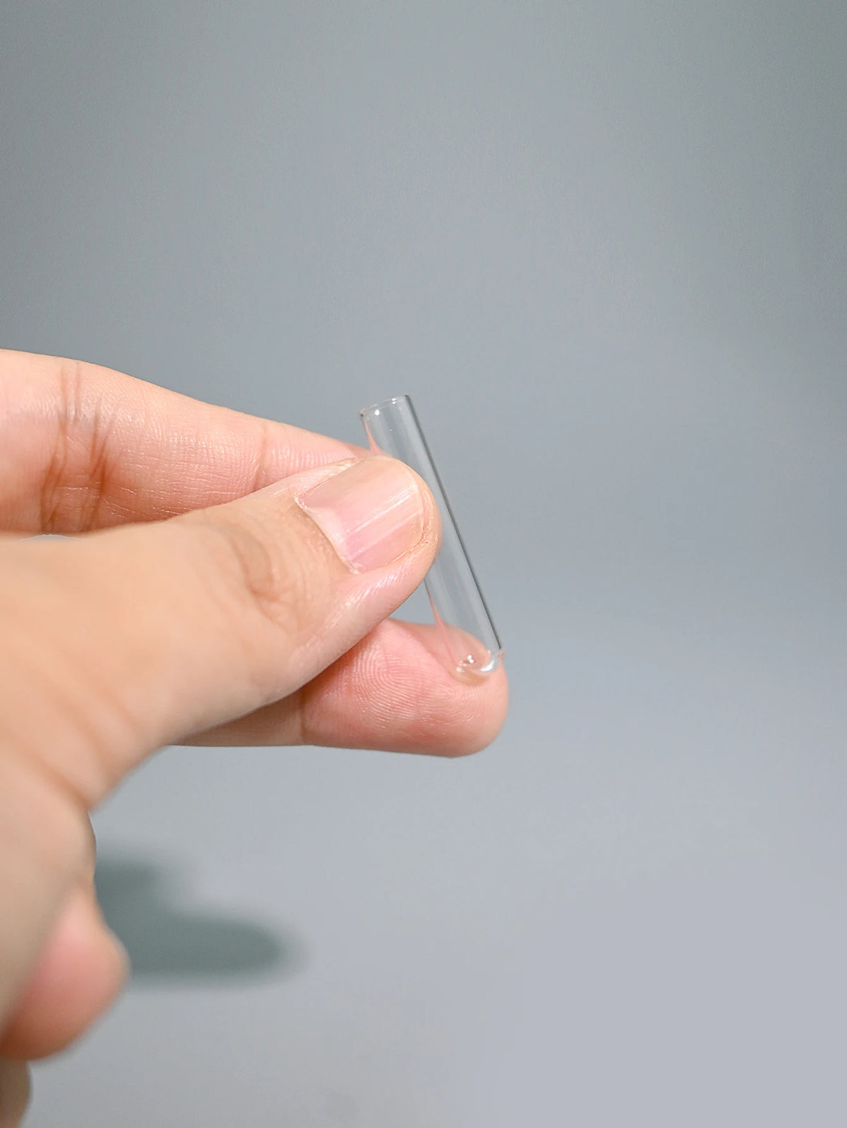 A hand holds a clear, thin glass tube or pipette against a light gray background.