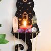 A black cat silhouette with a crescent moon, holding a lit candle and various crystals on a floating shelf.
