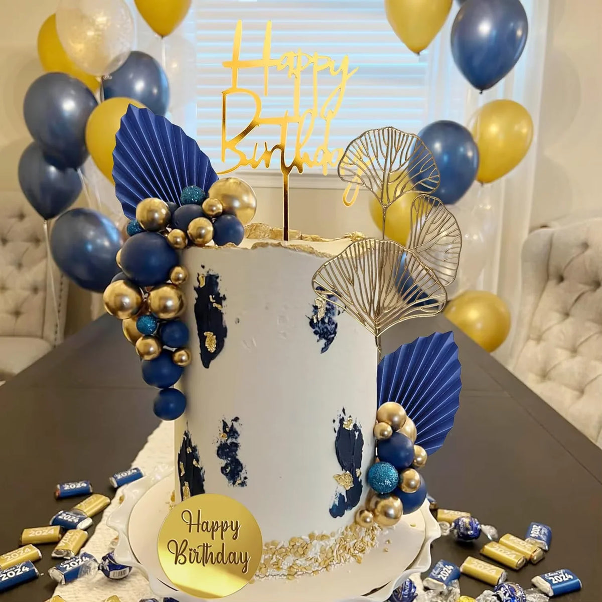 A white cake decorated with blue and gold paint splatters, surrounded by blue and gold balloons and foliage.