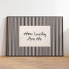 Black framed canvas wall art featuring the text How Lucky Are We on a white background with vertical black stripes.