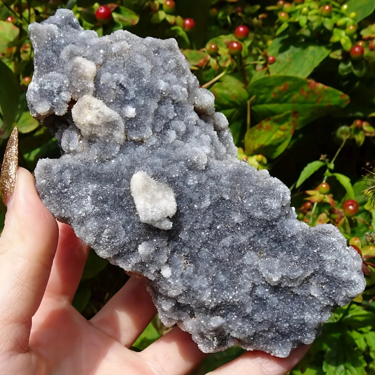 A hand holds a cluster of natural zinc crystals, appearing gray and translucent, against a green foliage background.