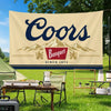 A beige flag banner with the word Coors in dark blue script is displayed outdoors on a grassy field.