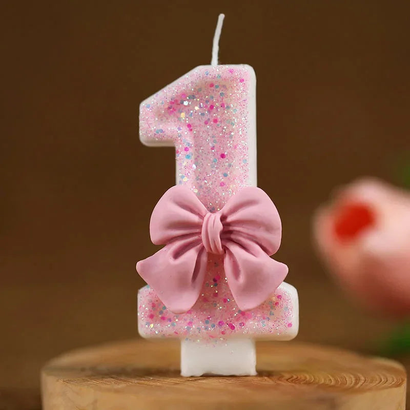 Una vela blanca con forma de número uno decorada con purpurina rosa y un gran lazo rosa.