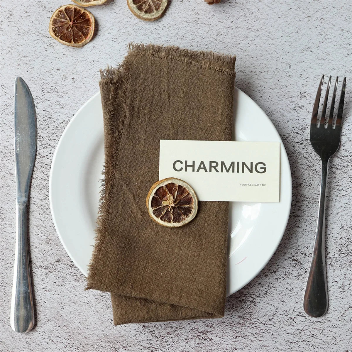 A set of handcrafted fringed cotton napkins is displayed on a white plate with a wooden fork and a slice of citrus fruit.