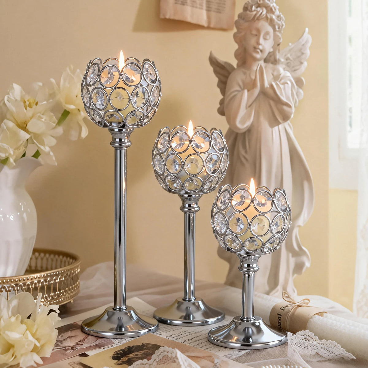 A silver metal glass candle holder stands next to a white flower arrangement and a statue of an angel.