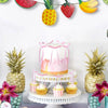 Pink and yellow coquette bow cake toppers displayed on a white cake stand with tropical fruit decorations.