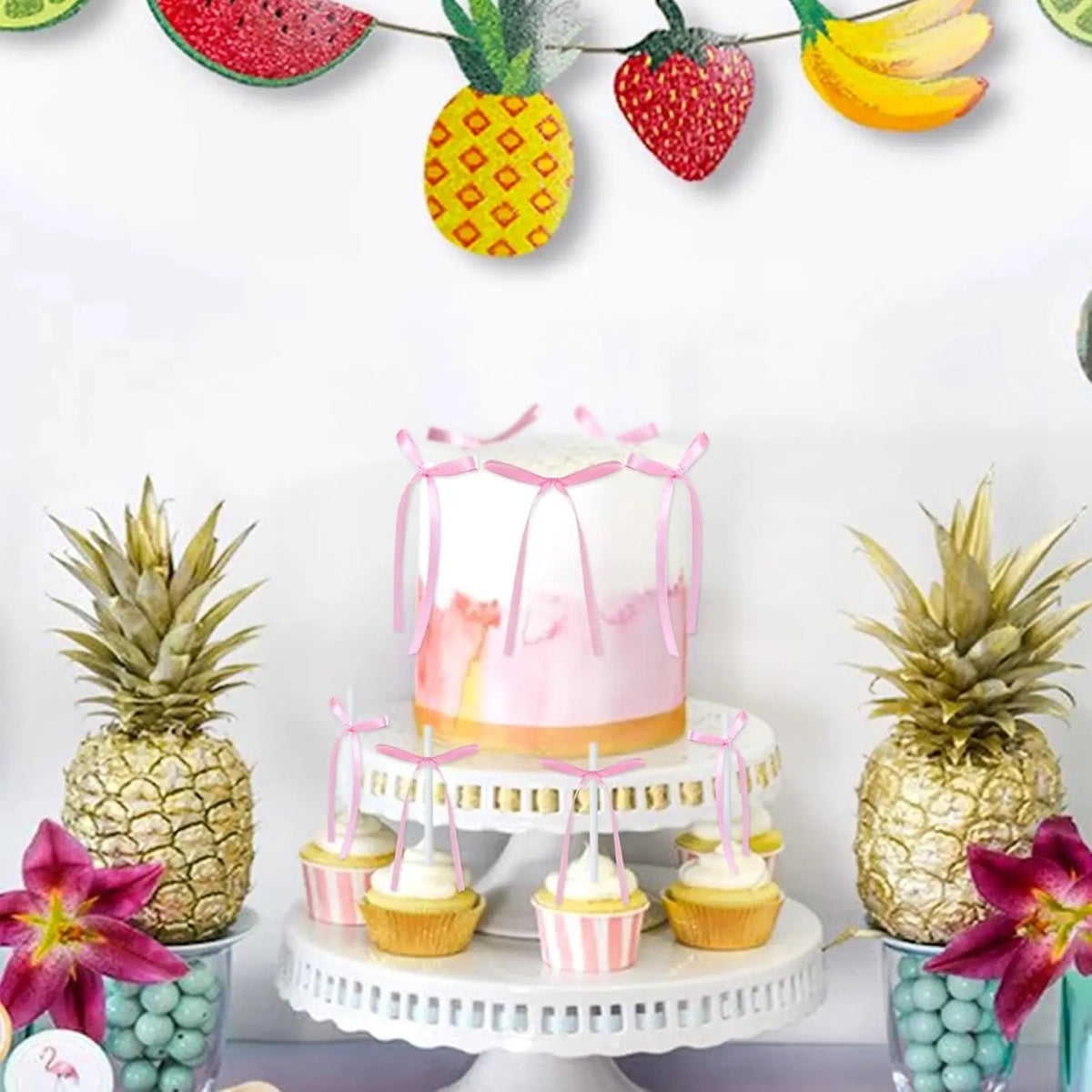 Pink and yellow coquette bow cake toppers displayed on a white cake stand with tropical fruit decorations.
