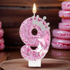 A pink glitter candle shaped like the number 9 with a crown, lit on a wooden surface.