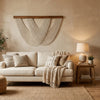 A bohemian macrame wall tapestry hangs above a light-colored sofa in a warmly lit living room.