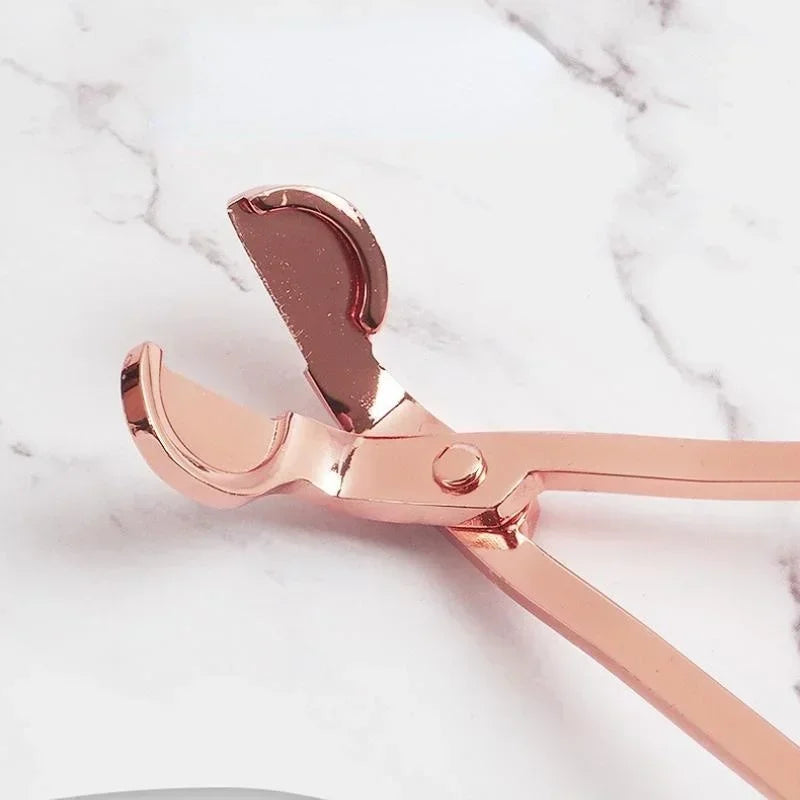 A pair of rose gold stainless steel candle wick trimmer scissors resting on a white marble surface.