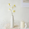 A white ceramic vase holds several white daisies on a textured surface.