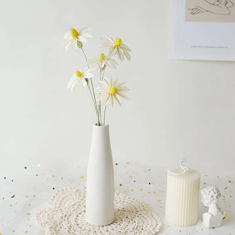 A white ceramic vase holds several white daisies on a textured surface.
