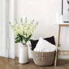 A white artificial delphinium bouquet in a ceramic vase sits on a wooden table next to a black cushion.