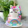 A tiered white and pink unicorn cake decorated with colorful flowers, stars, and shiny balls on a rainbow base.