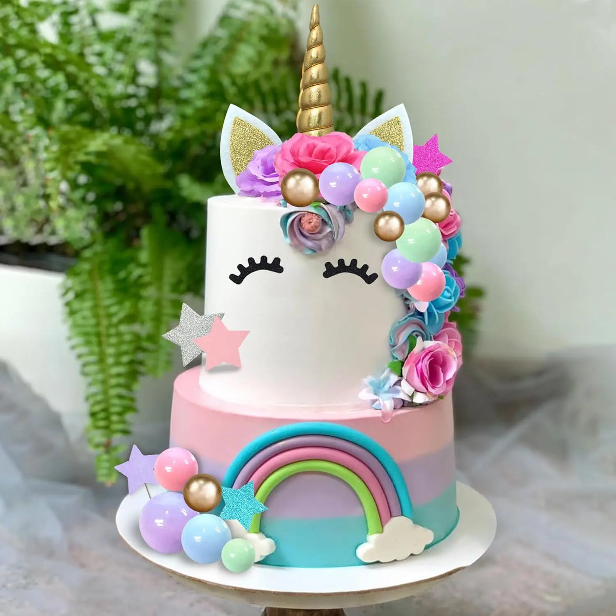 A tiered white and pink unicorn cake decorated with colorful flowers, stars, and shiny balls on a rainbow base.