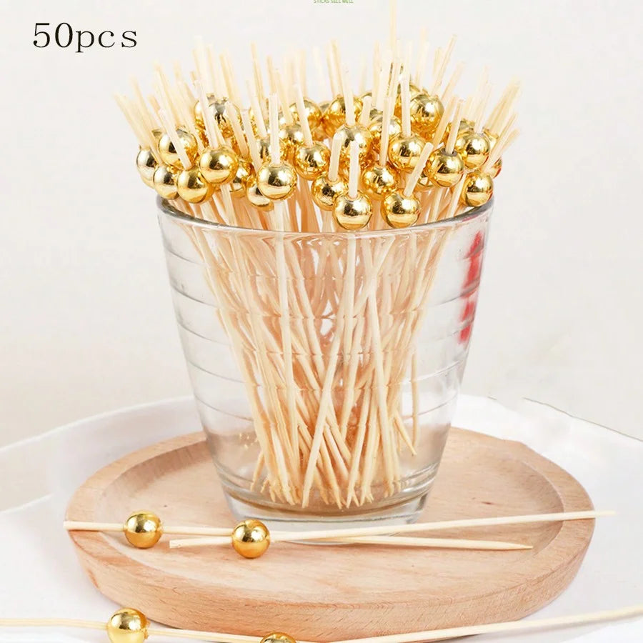 A glass bowl filled with 50 bamboo fruit skewers topped with gold-colored spherical beads.
