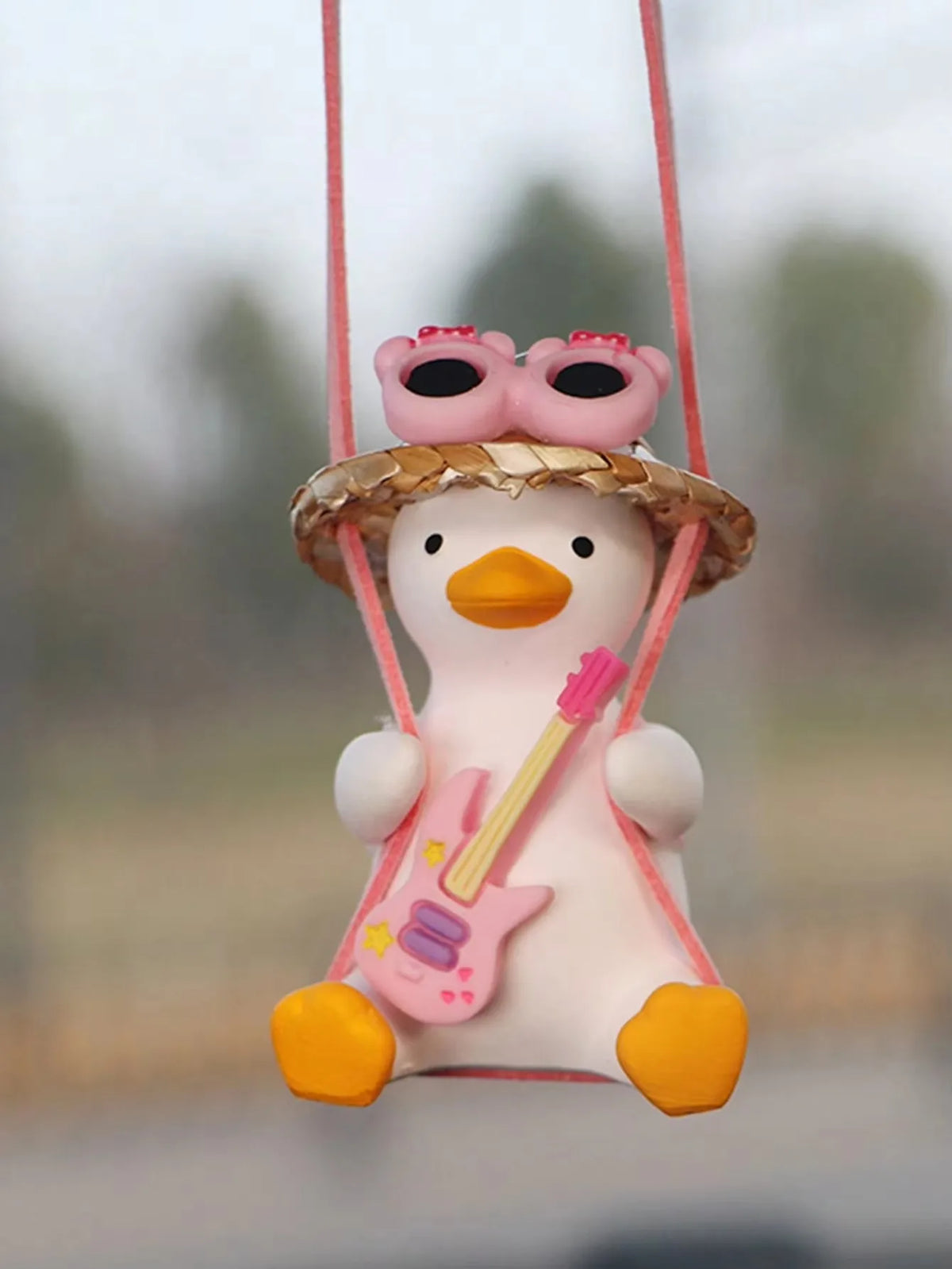 A cute pink anime duck ornament wearing a straw hat and sunglasses is suspended by a pink string.