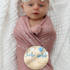 A newborn birth announcement sign featuring a baby wrapped in pink fabric with white text and a cloud-shaped disk.