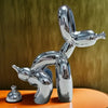 A chrome sculpture of a dog squatting, displayed on an orange surface against a muted green background.