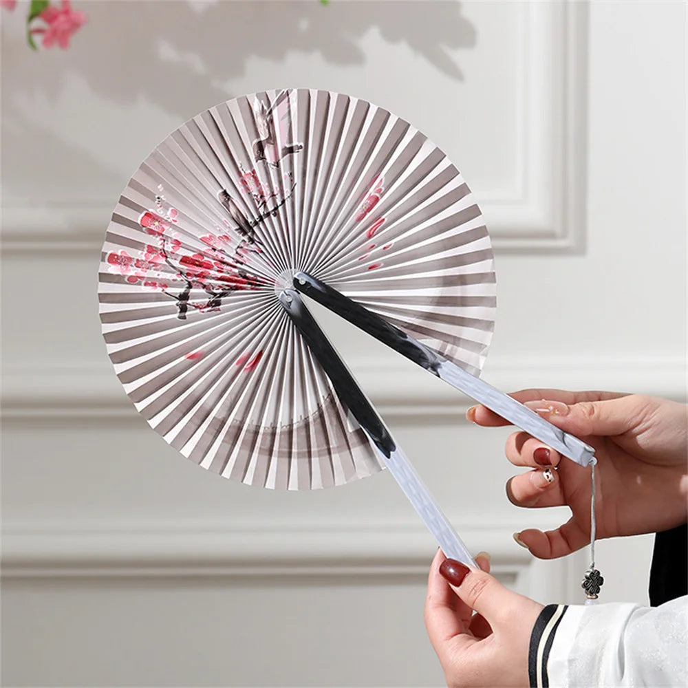 A traditional folding fan with pink floral designs and a tassel is held by a person.