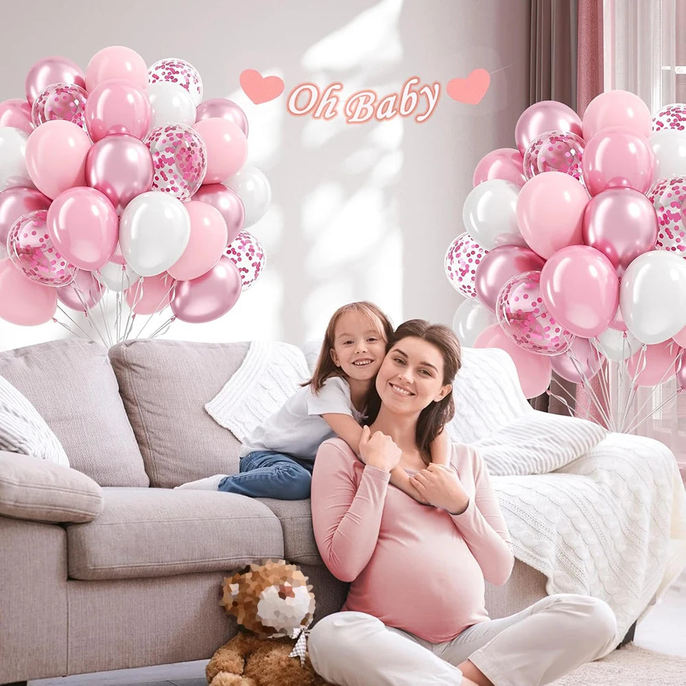 A pregnant woman sits on a sofa surrounded by pink and white latex balloons with confetti accents.