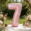 A large pink foil number balloon shaped like the number seven stands on a white table surrounded by floral arrangements and a