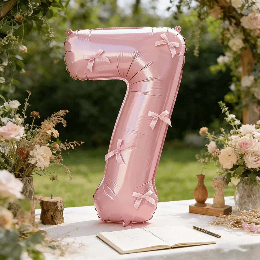 A large pink foil number balloon shaped like the number seven stands on a white table surrounded by floral arrangements and a