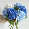 A hand holds a bouquet of blue and white hydrangea silk flowers with green stems.