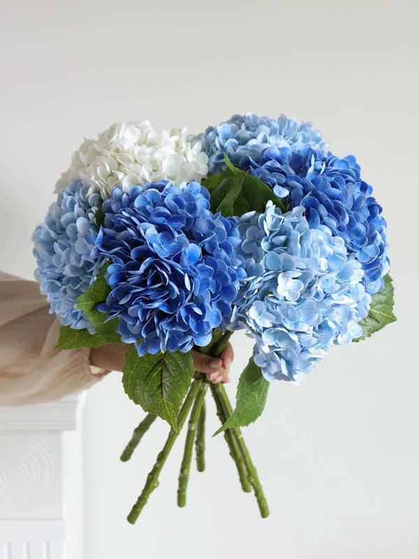 A hand holds a bouquet of blue and white hydrangea silk flowers with green stems.
