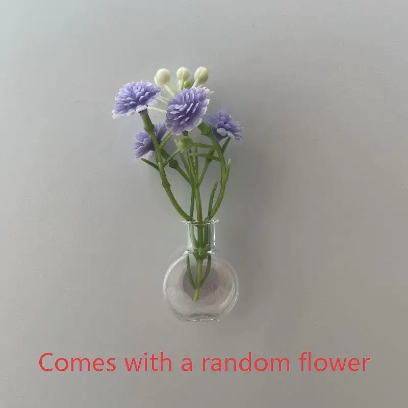 A small glass vase holds a sprig of purple and white flowers on a light gray background.