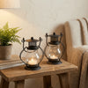 A decorative metal candlestick lantern with a glass bulb and lit candle sits on a wooden table next to a potted plant.