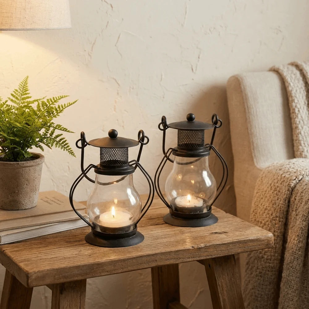 A decorative metal candlestick lantern with a glass bulb and lit candle sits on a wooden table next to a potted plant.