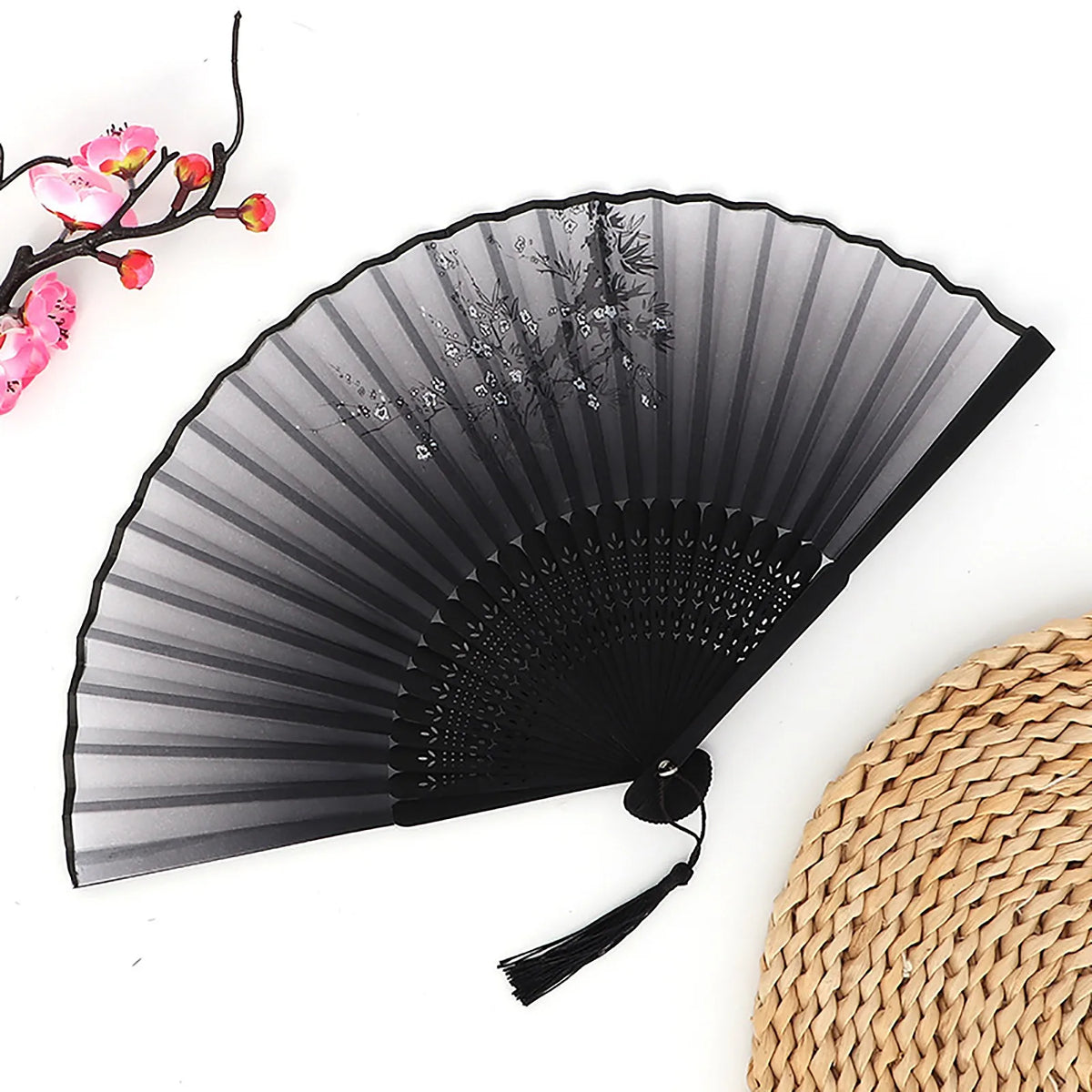 A black vintage bamboo folding fan with sheer fabric and pink floral accents is shown next to a woven straw mat.