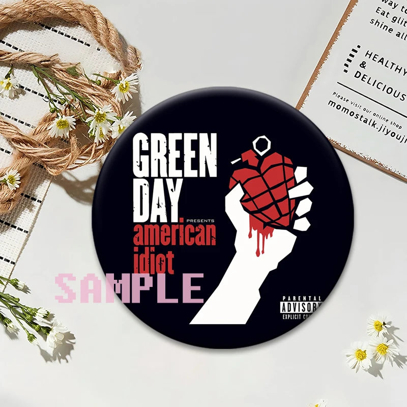 A black button pin featuring a hand holding a red, textured object with the text GREEN DAY PRESENTS american idiot SAMPLE