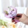 Une main tient un bouquet de roses artificielles violettes et de fleurs de remplissage blanches dans du papier rose.