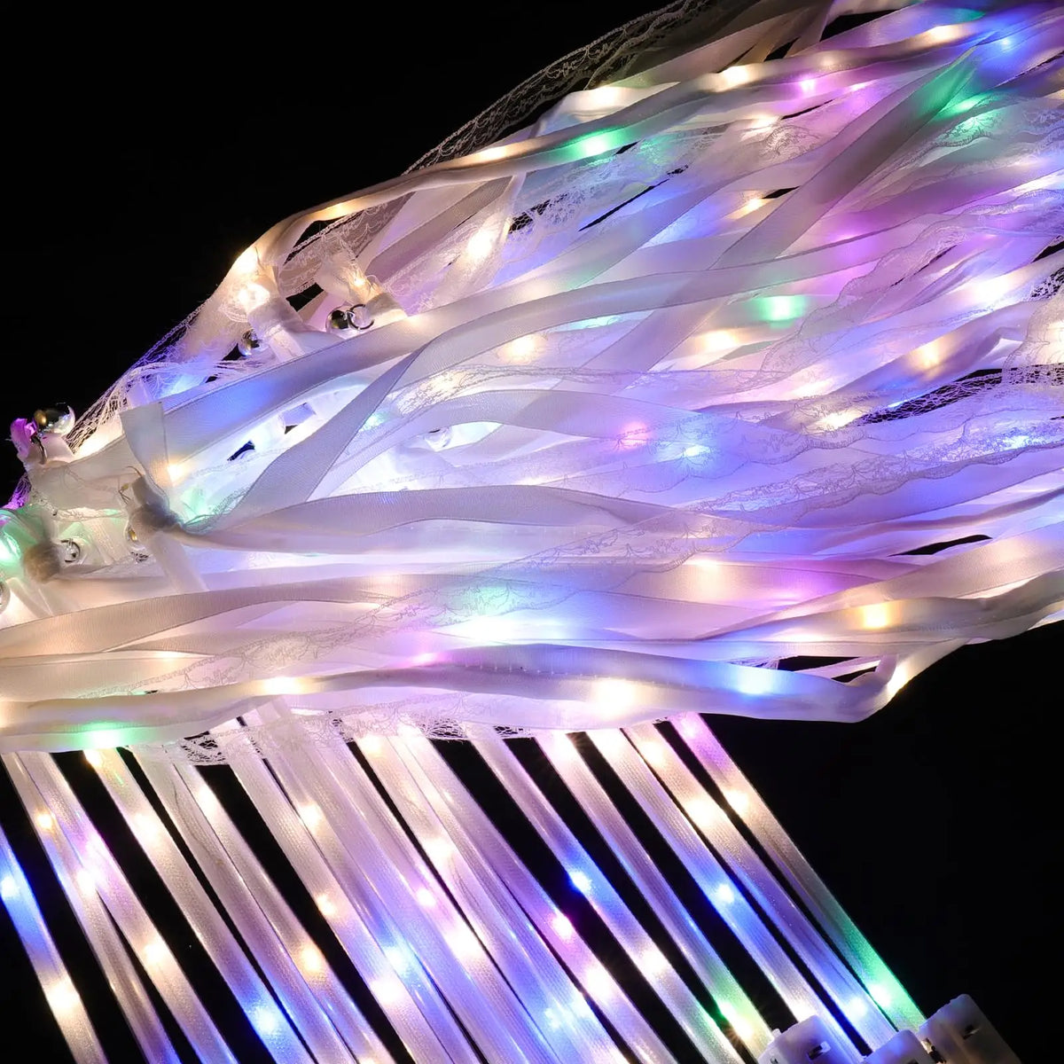 A close-up of LED glow wands with ribbons and bells illuminating a soft purple and green light against a dark background.