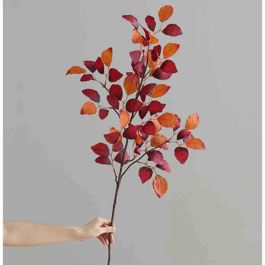 A hand holds a branch of autumn beech simulation leaves with reddish-orange and deep red colors against a gray background.