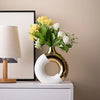 A golden ceramic flower vase filled with yellow and white flowers sits on a white cabinet next to a lamp.