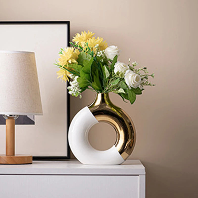 A golden ceramic flower vase filled with yellow and white flowers sits on a white cabinet next to a lamp.