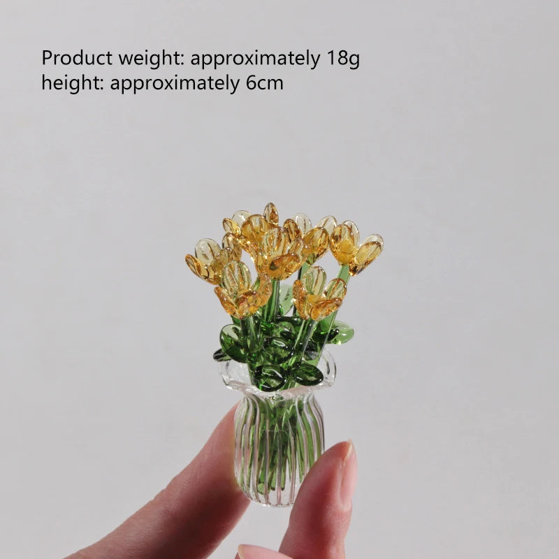 A hand holds a small bouquet of yellow glass flowers in a clear glass vase.