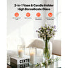 A clear glass cylinder vase with a pillar candle is displayed on a glass table next to a bouquet of white flowers.