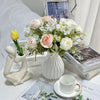 A white ceramic flower vase filled with white and pink flowers sits next to a white sculpture.