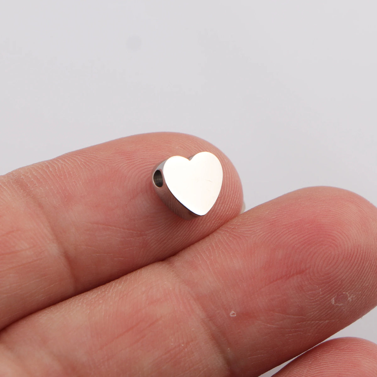 A silver heart-shaped stainless steel bead charm is held between fingers.