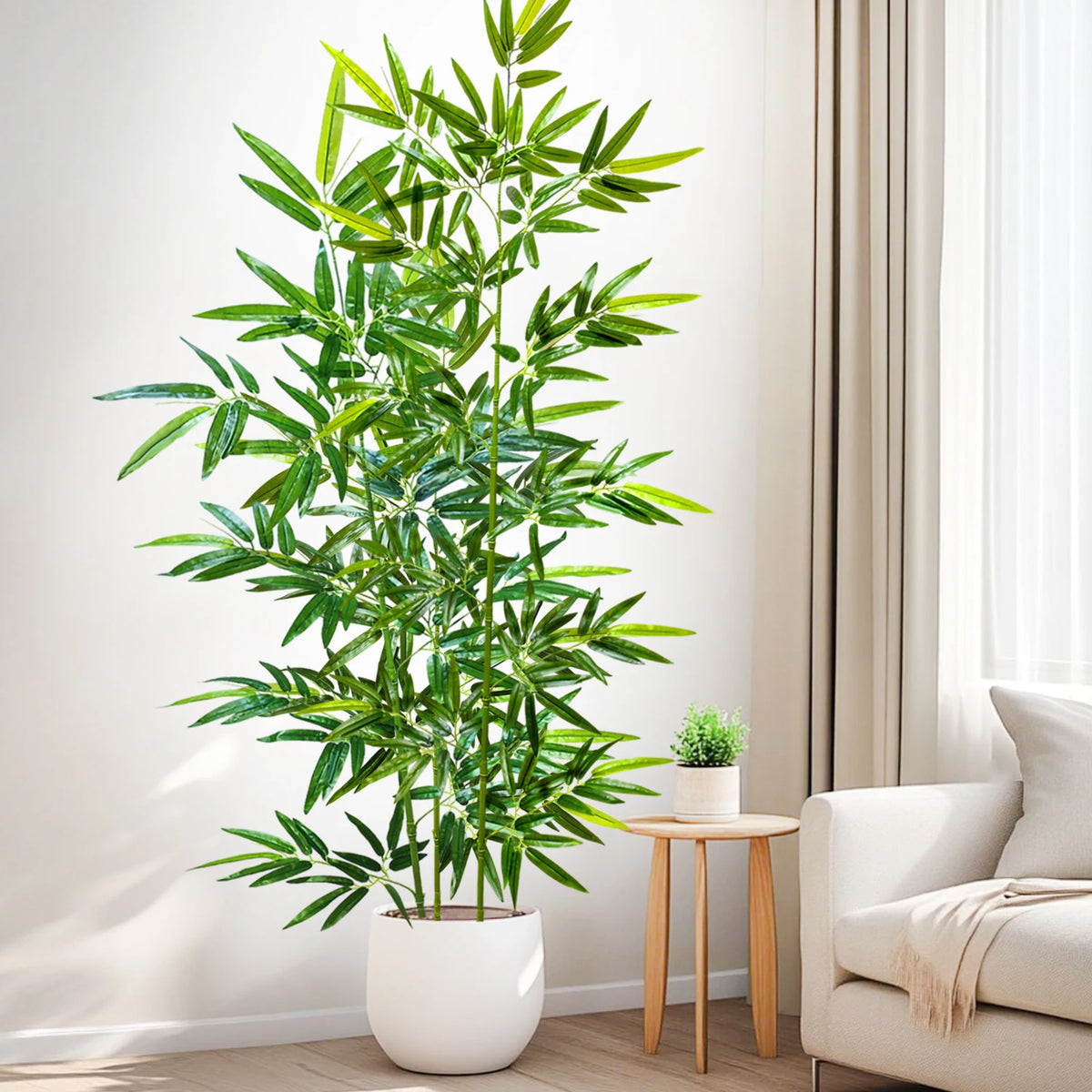 An artificial bamboo branch with green leaves displayed in a white pot on a wooden table next to a light-colored sofa.