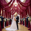 A wedding ceremony featuring a bride and groom standing in front of a luxurious magenta draped backdrop with chandeliers.